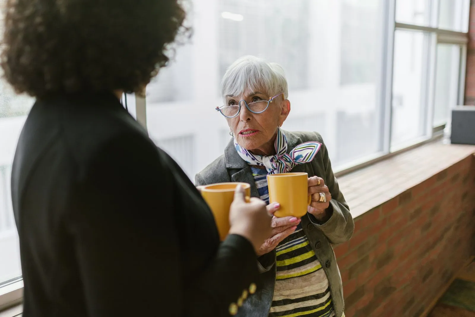 Elderly woman conversing with a colleague while holding coffee mugs indoors.